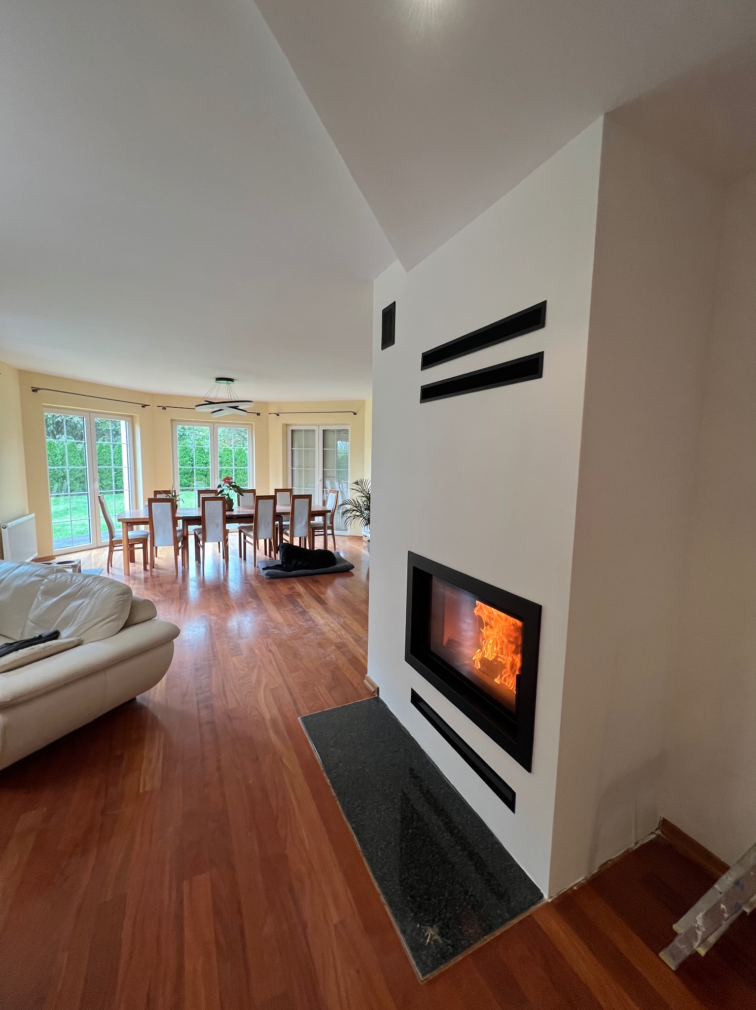 Nowoczesny wkład kominkowy z czarną ramą, wbudowany w białą ścianę z poziomymi, czarnymi elementami dekoracyjnymi, widoczne płomienie, w tle salon z drewnianą podłogą, stołem i krzesłami.