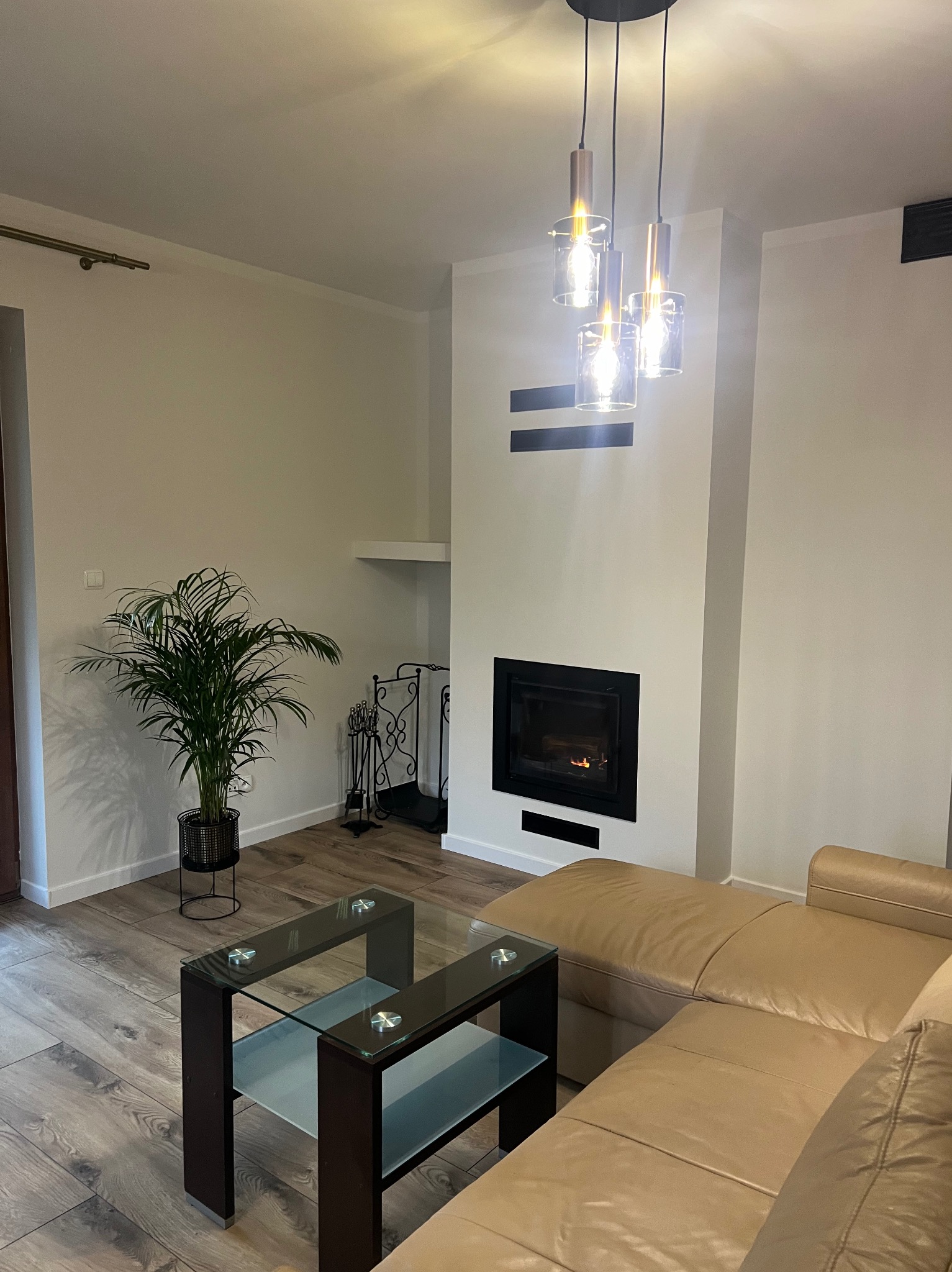 Nowoczesny salon z wkładem kominkowym w zabudowie, szklanym stolikiem kawowym i beżową sofą narożną, oświetlony wiszącymi lampami.