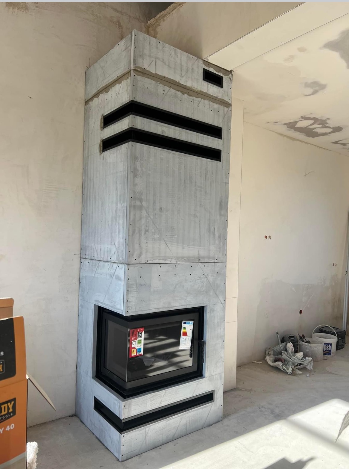 Surowy, szary obudowany wkład kominkowy z czarnymi poziomymi akcentami w trakcie montażu, widoczne niedokończone ściany i narzędzia budowlane na podłodze.