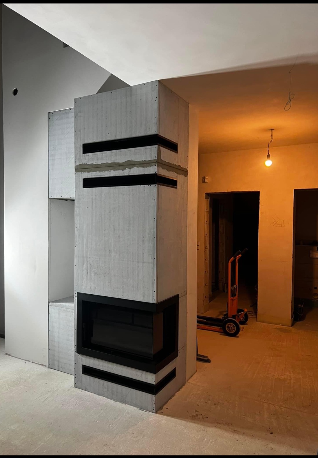 Surowy, betonowy wkład kominkowy z czarną ramą, w trakcie montażu; widoczne niedoskonałości wykończenia i wózek transportowy w tle.