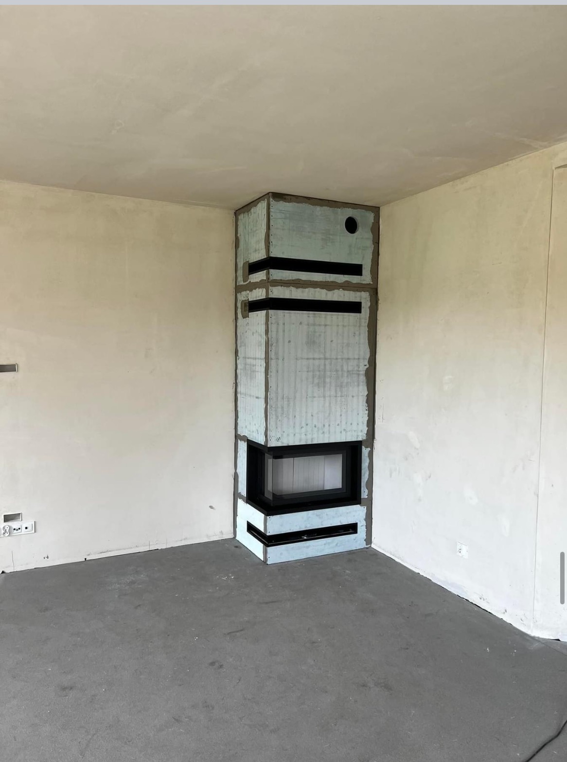 Narożny kominek w trakcie obudowy, widoczne surowe wykończenie z betonu architektonicznego z czarnymi wstawkami, ściany i podłoga w stanie deweloperskim.