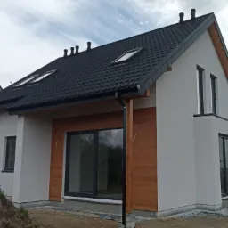 Nowoczesny dom jednorodzinny z szarą elewacją, drewnianymi akcentami i ciemną dachówką, widoczne okna dachowe i rynny