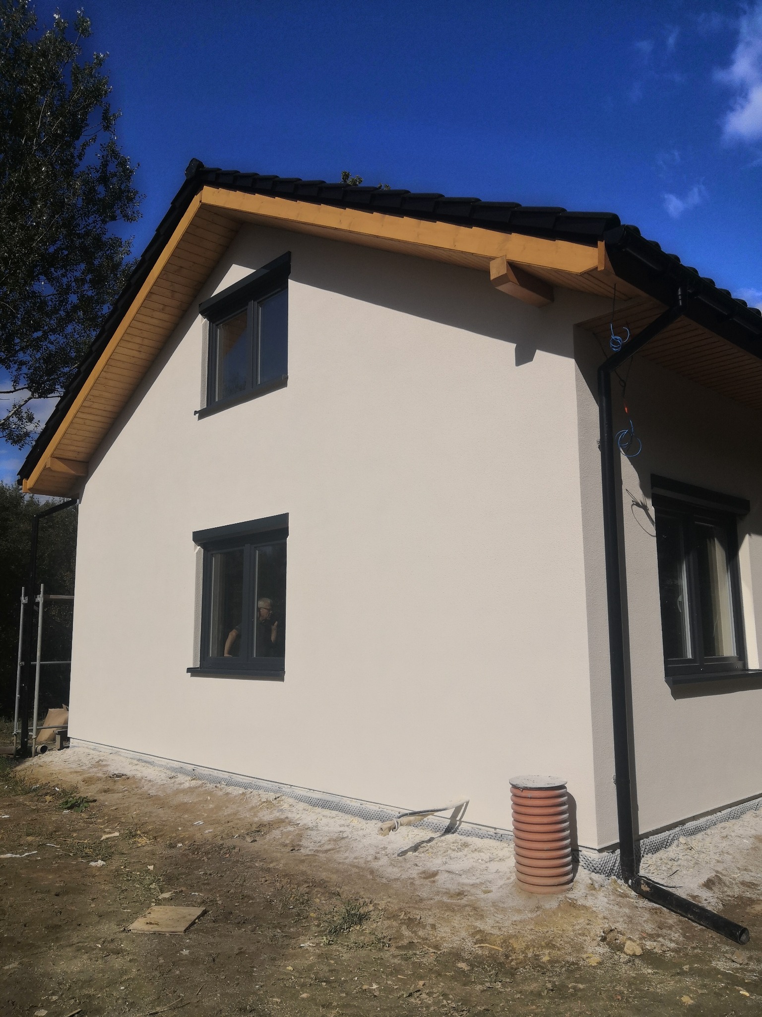 Dwukondygnacyjny dom z jasną elewacją, ciemną stolarką okienną i czarnym dachem, widoczny z boku, z osobą wyglądającą przez okno na parterze.