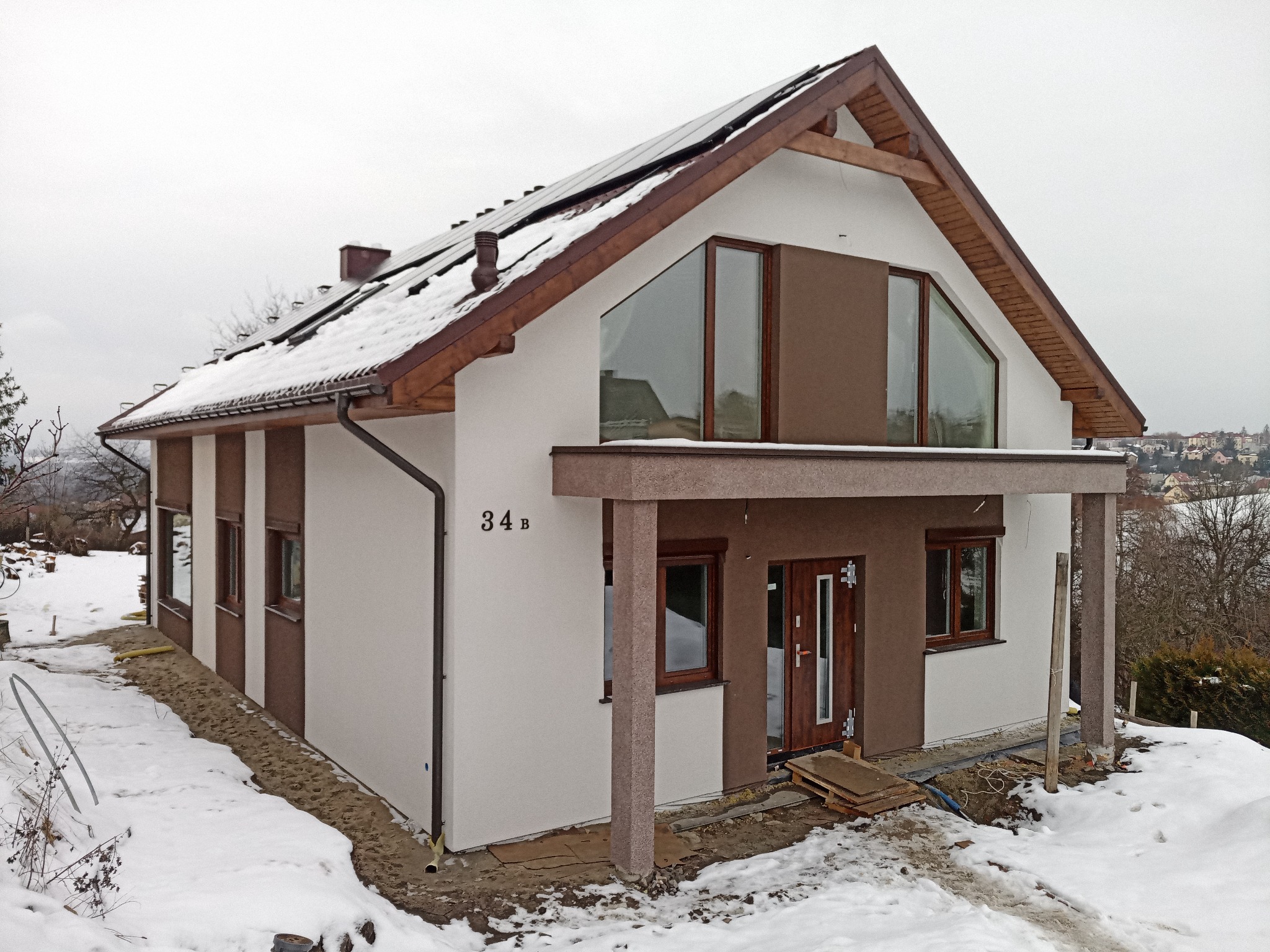 Dwukondygnacyjny dom z białymi ścianami i brązowymi akcentami architektonicznymi, z panelami słonecznymi na dachu pokrytym śniegiem, widoczny numer domu '34 B' oraz teren wokół w trakcie prac...