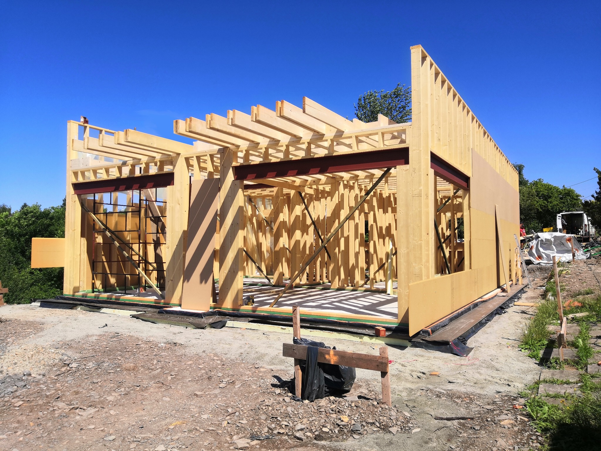 Wznoszenie szkieletu drewnianego domu z widocznymi elementami konstrukcyjnymi, w tym belkami stropowymi i stalowymi wzmocnieniami, na przygotowanym fundamencie z izolacją, w otoczeniu placu budowy...