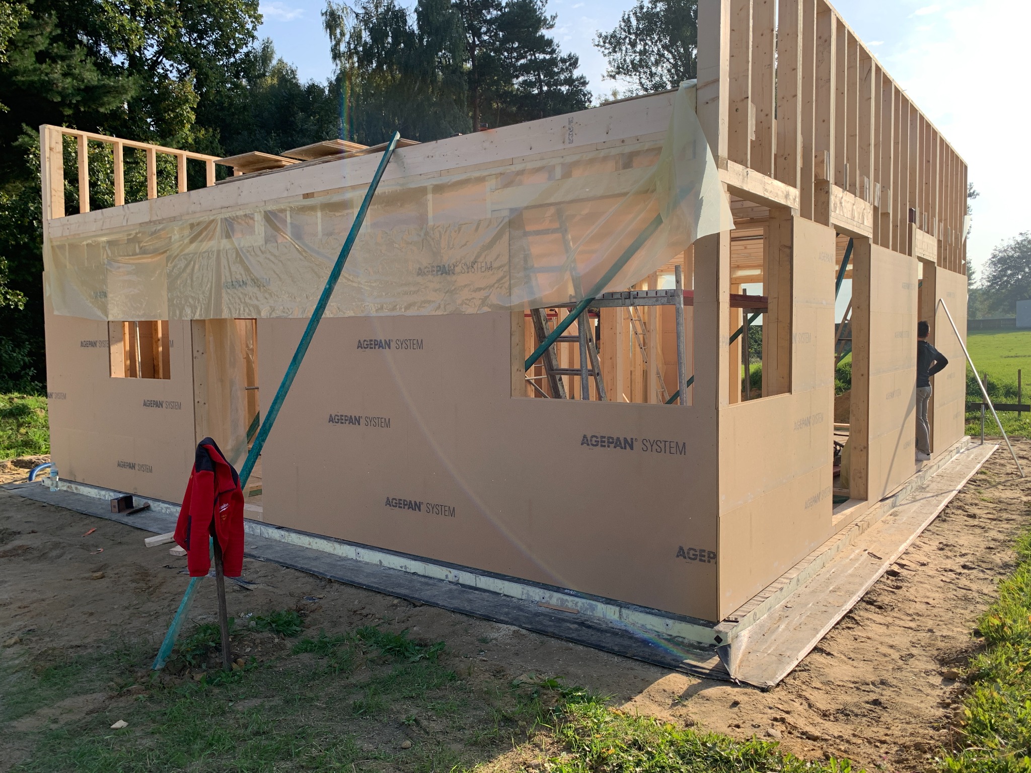 Wznoszenie konstrukcji domu z drewnianym szkieletem, widoczne płyty AGEPAN SYSTEM, ocieplenie i elementy konstrukcyjne, czerwona kurtka na tyczce w pierwszym planie.