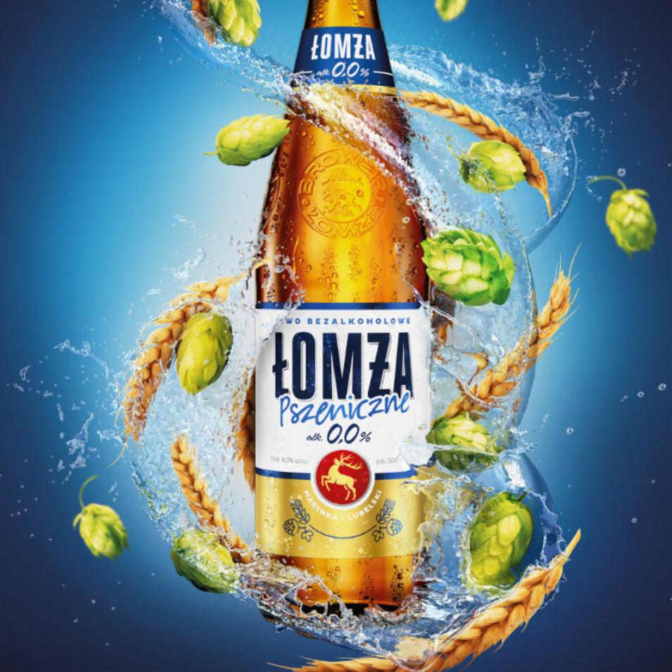 Butelka bezalkoholowego piwa Łomża Pszeniczne 0.0% w dynamicznym ujęciu, otoczona chmielem i jęczmieniem w rozbryzgach wody na niebieskim tle.