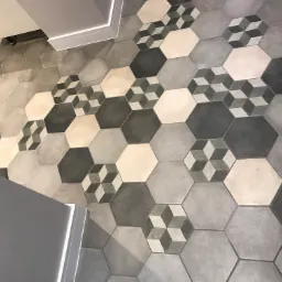 Ułożona pod kątem podłoga z heksagonalnych płytek w odcieniach szarości, beżu i czerni, niektóre z wzorem 3D, widok na fragment ściany z listwą przypodłogową.