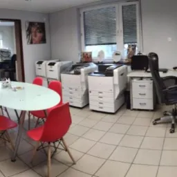 Panoramiczny widok biura z kilkoma drukarkami wielofunkcyjnymi, okrągłym stołem z czerwonymi krzesłami, półkami z ceramiką artystyczną i plakatem motocykla na ścianie.