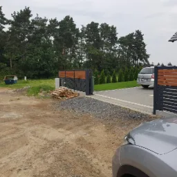 Nowoczesna brama wjazdowa z poziomymi panelami z ciemnego metalu i drewnopodobnymi wstawkami, widok na podjazd wyłożony szarą kostką brukową i fragment domu z szarym dachem