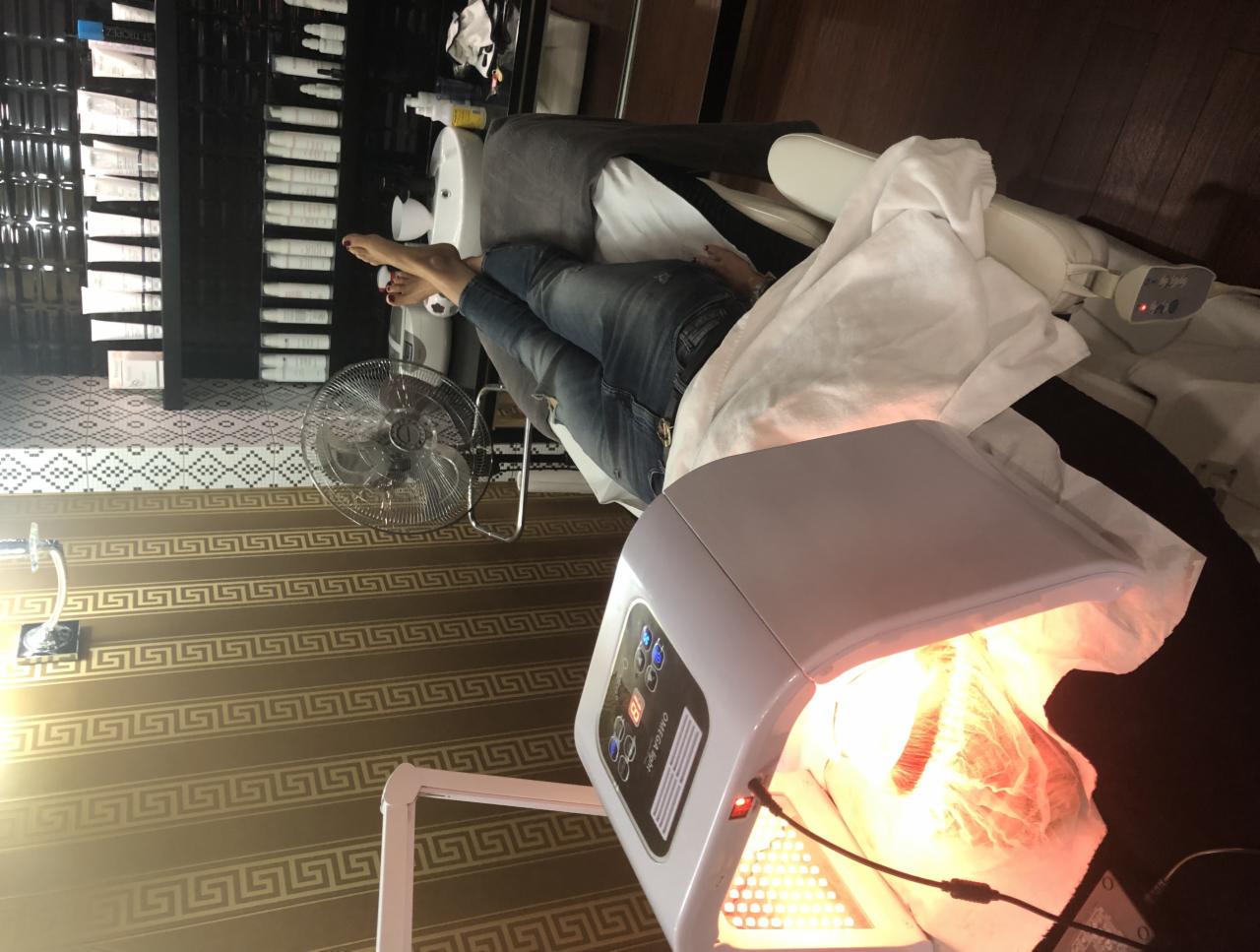 Klientka leży na leżance pod lampą LED w gabinecie, widoczne kosmetyki na półce w tle.