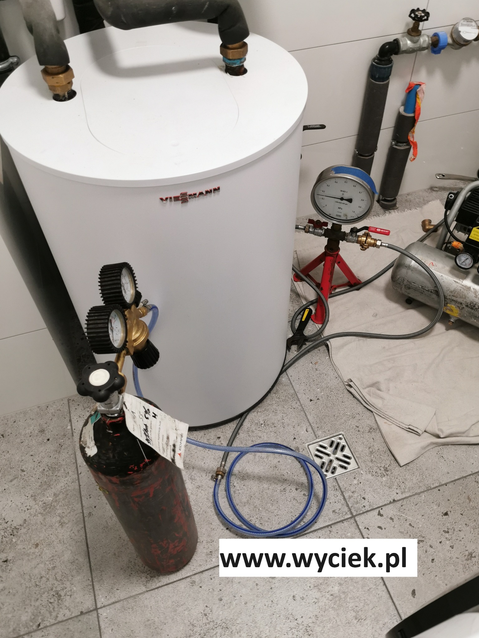 Biały zbiornik Viessmann podłączony do butli z gazem i manometru, używany do lokalizacji wycieków. Widoczny kompresor i kratka ściekowa w pomieszczeniu z białymi płytkami.