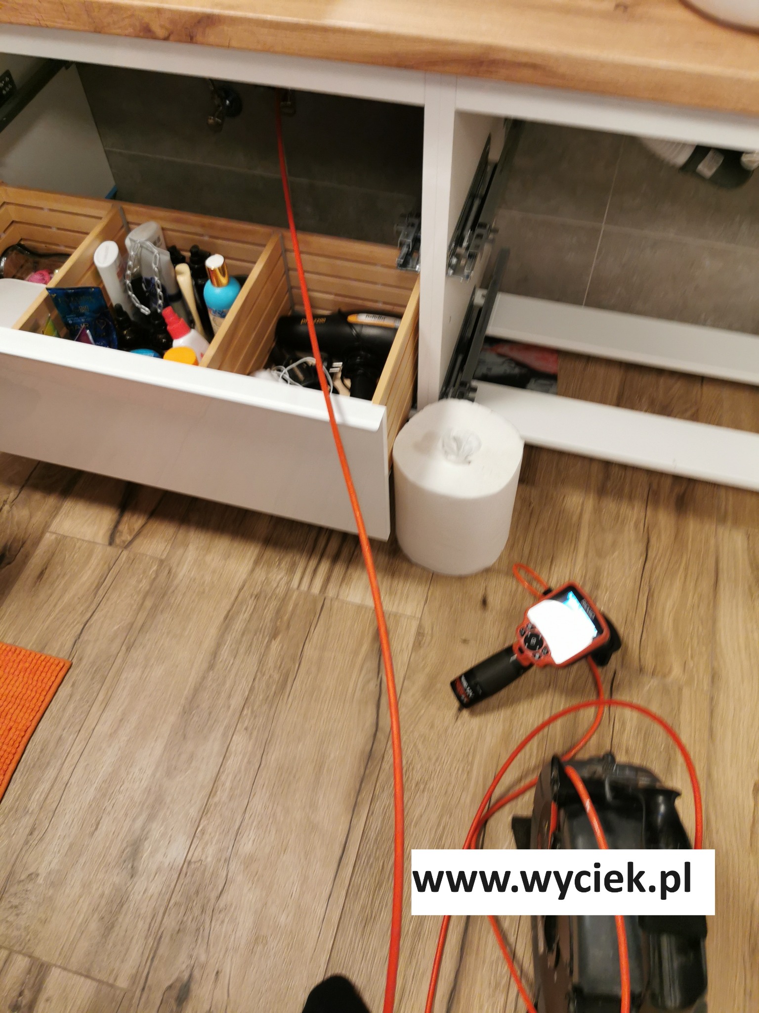 Inspekcja endoskopowa pod szafką z otwartą szufladą, widoczny sprzęt diagnostyczny i przewód wprowadzony w głąb. Parkiet imitujący drewno w tle.