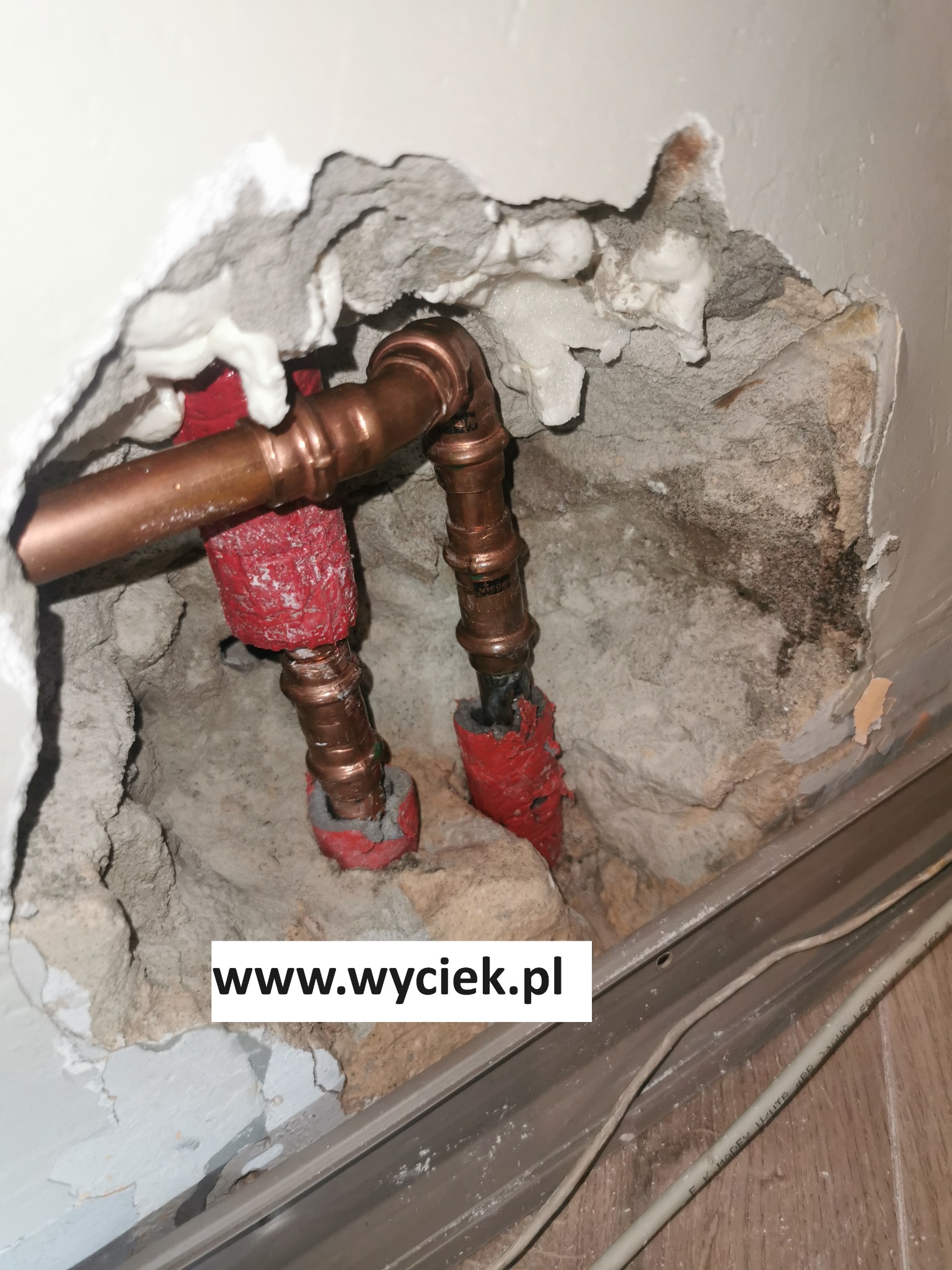Odsłonięte miedziane rury w ścianie z ubytkiem tynku, zabezpieczone czerwoną izolacją. Widoczne ślady wilgoci i pianka montażowa. Ujęcie z dołu, blisko podłogi.