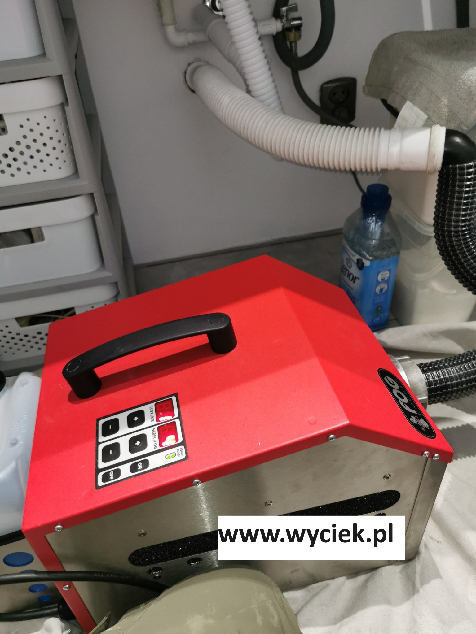 Czerwone urządzenie FOG z wężem pod zlewem, z widocznymi rurami i butelką płynu. Sprzęt do usuwania wilgoci w trudno dostępnych miejscach.