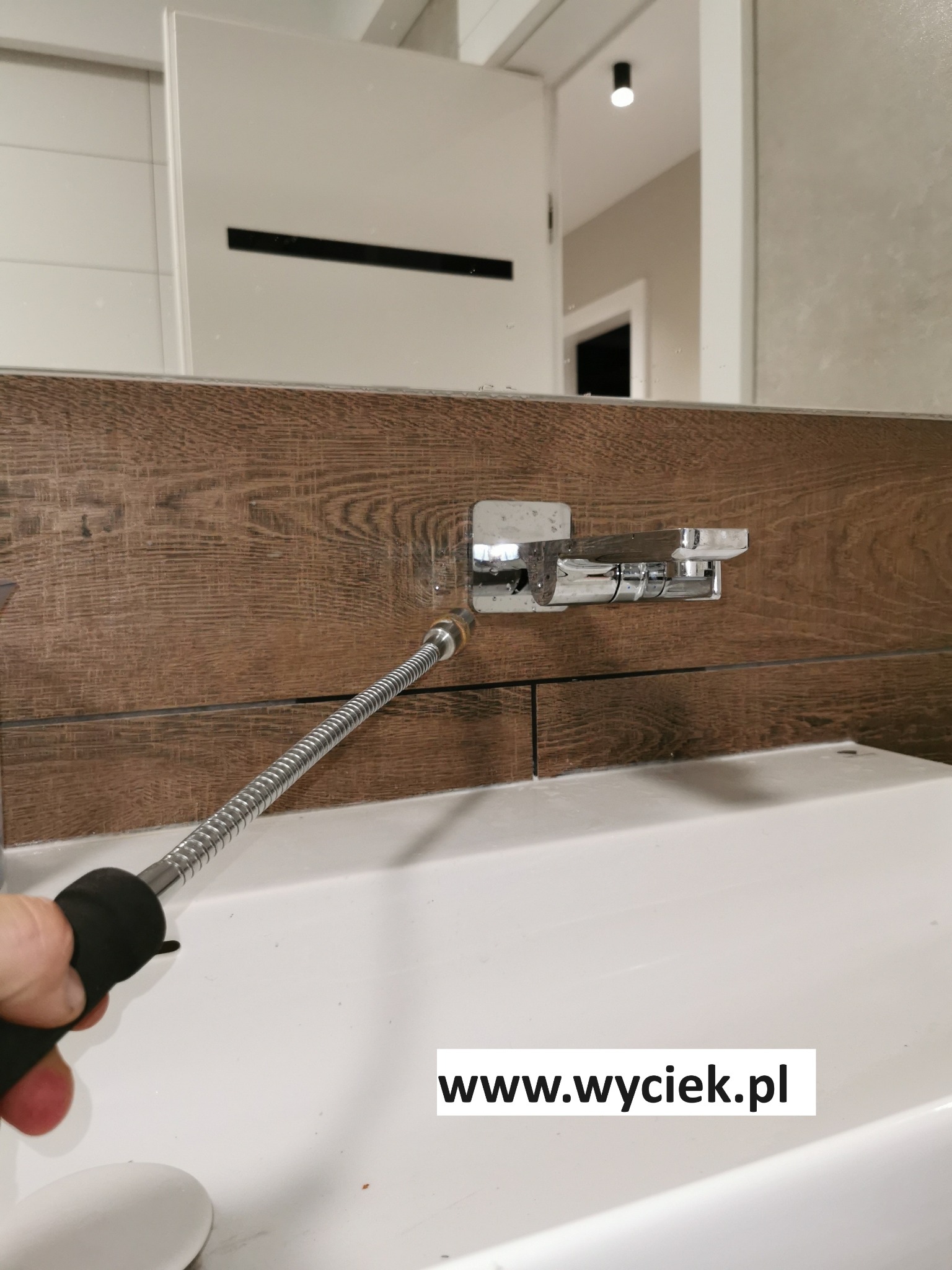 Sprawdzanie szczelności baterii umywalkowej elastycznym wężem. Widoczne płytki imitujące drewno i biała umywalka. Detekcja wycieków w łazience.