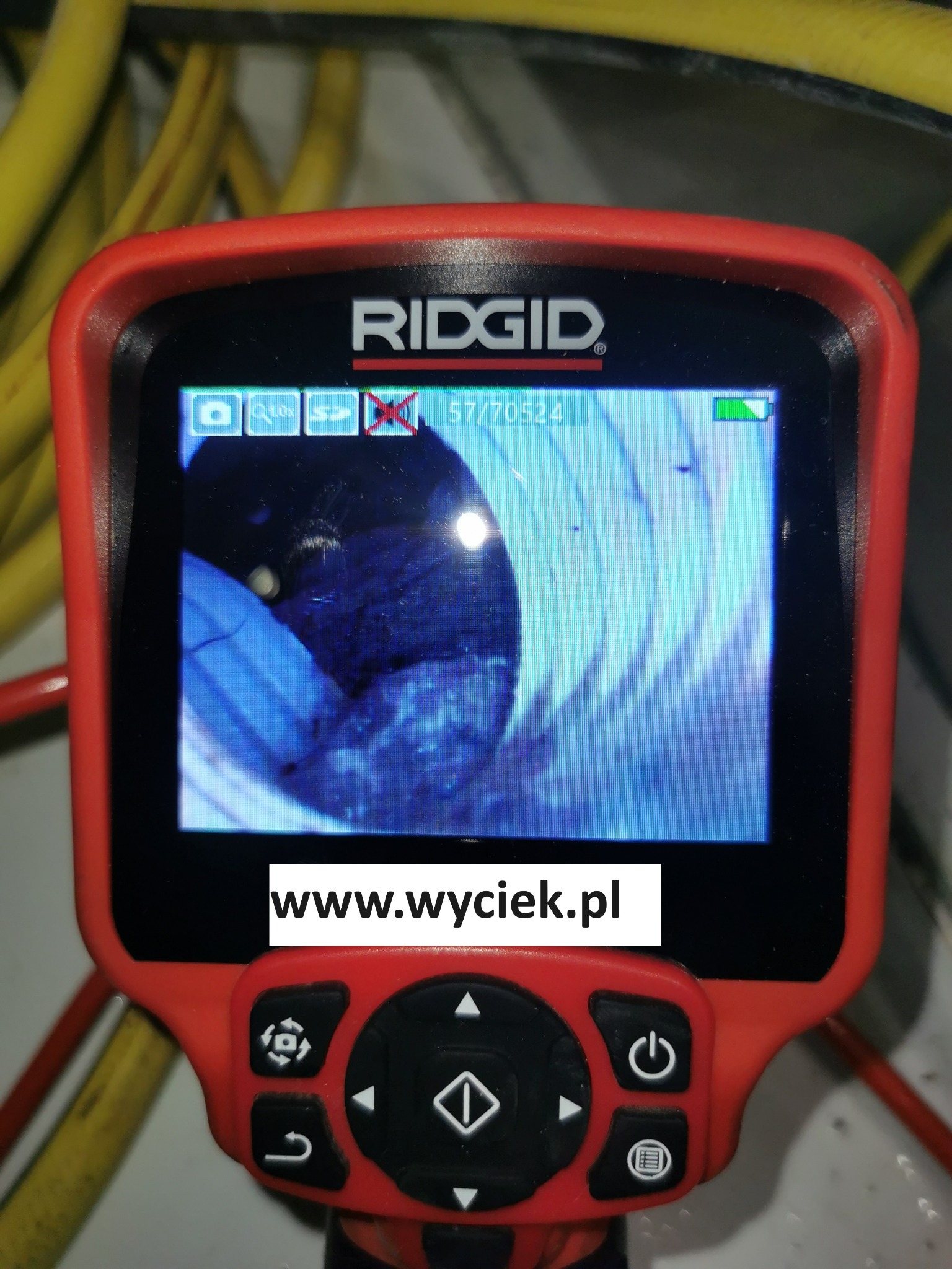 Kamera inspekcyjna RIDGID z widokiem wnętrza rury, prawdopodobnie kanalizacyjnej, na ekranie. Widoczny adres strony www.wyciek.pl. Żółty kabel w tle.