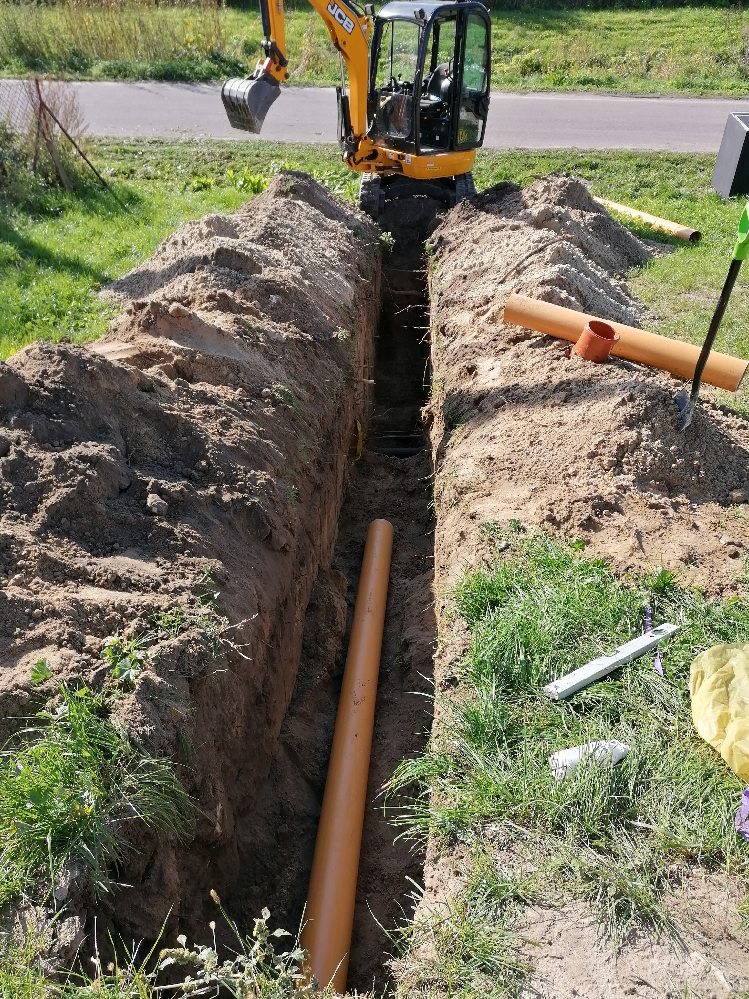 Wykop pod rury kanalizacyjne z pomarańczowymi rurami, koparka JCB w tle, widoczny poziomica i łopata.