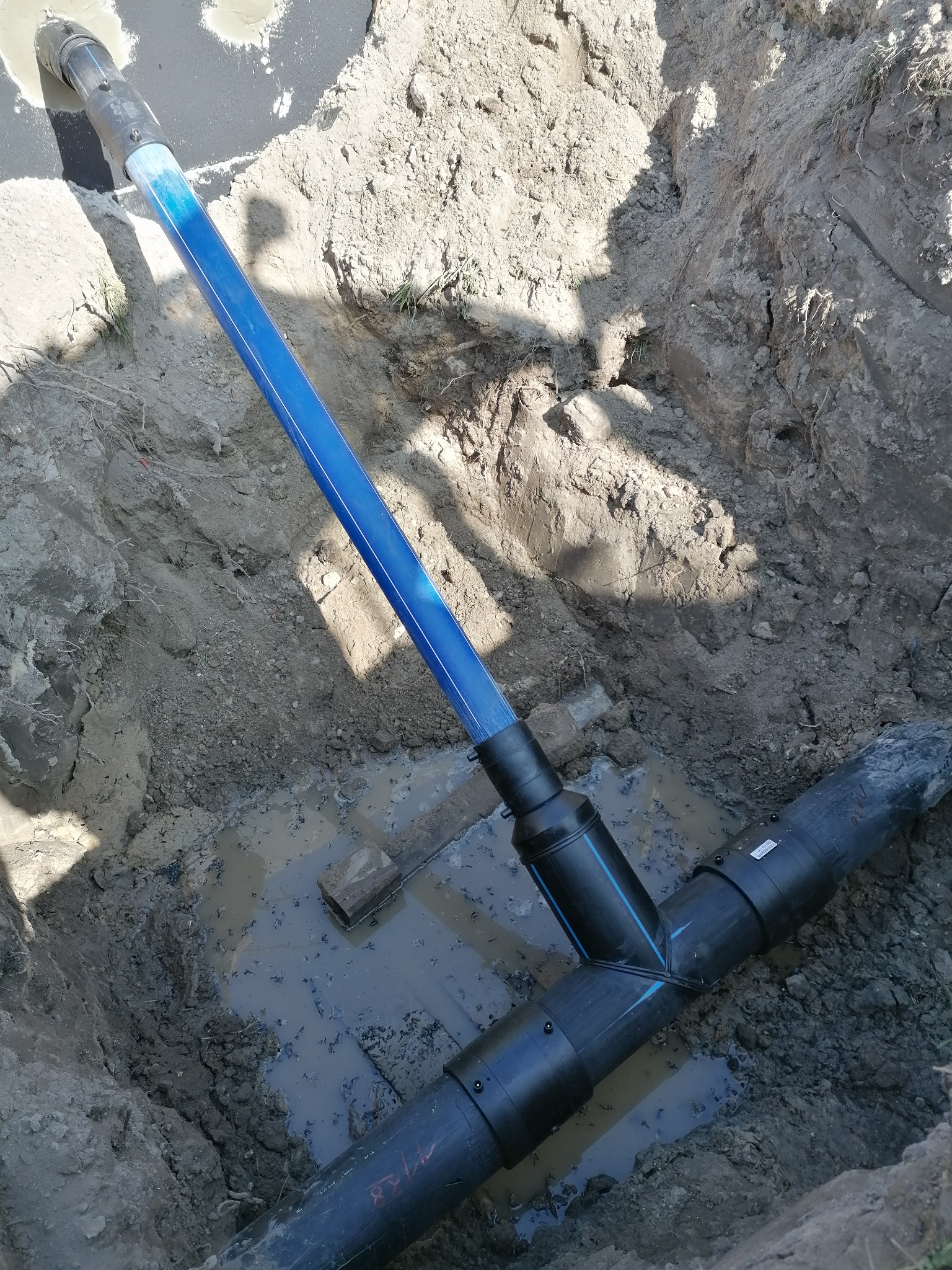 Wykop z widocznymi rurami wodociągowymi z niebieskim paskiem, połączonymi trójnikiem, częściowo zalany wodą.