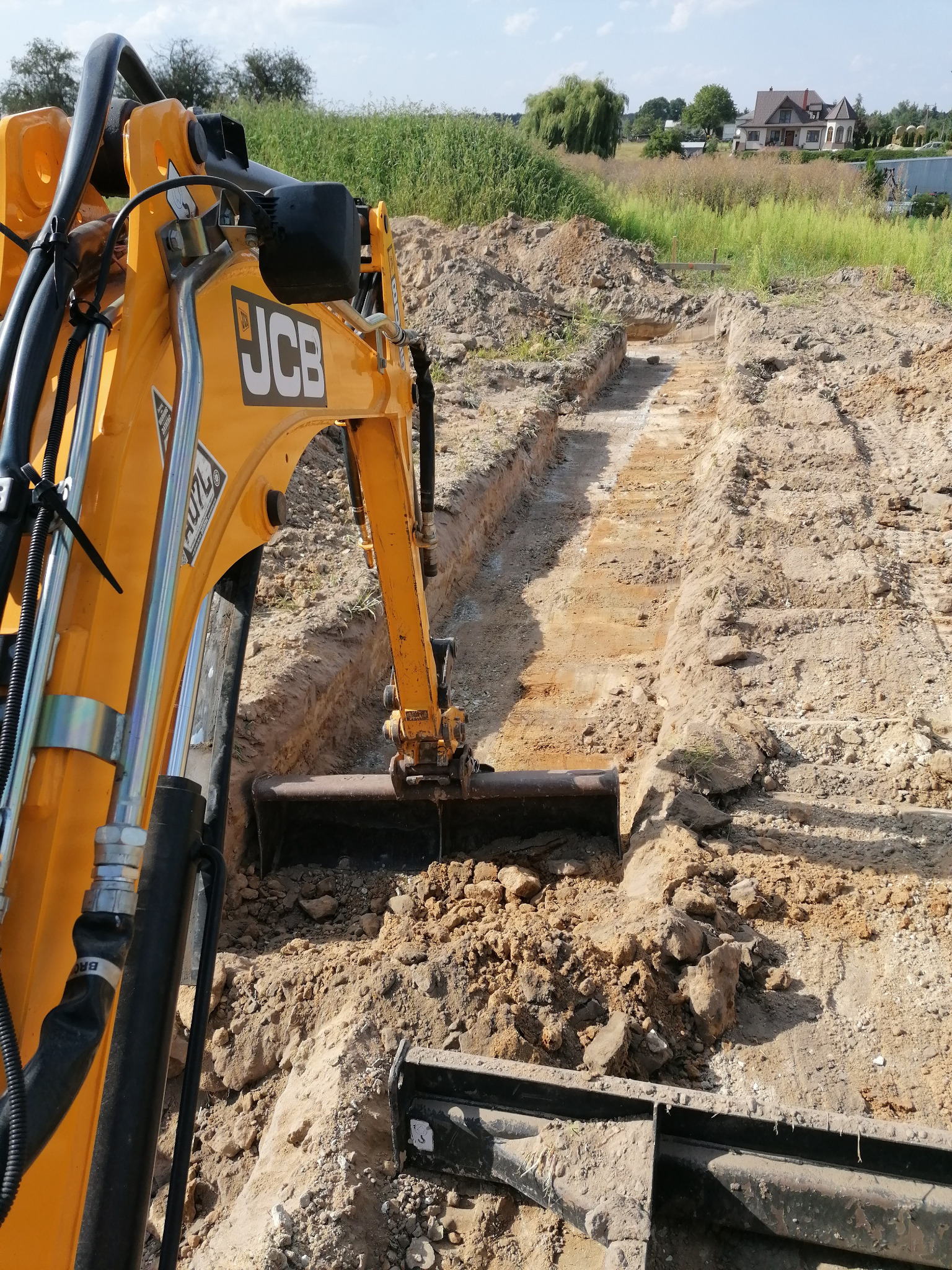 Żółta koparka JCB wykopuje wąski rów w ziemi, prawdopodobnie pod instalację, w tle widoczne zabudowania mieszkalne i zieleń.