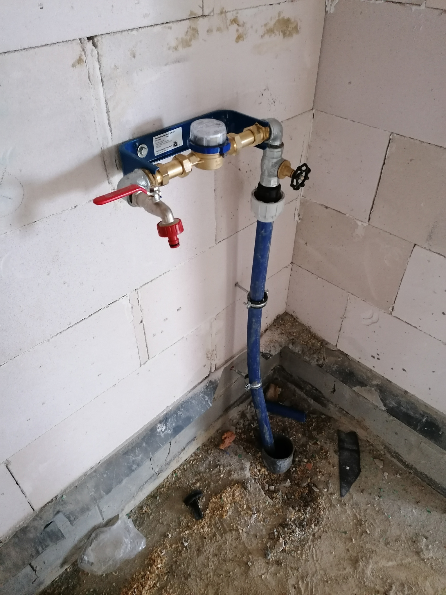 Zmontowany zestaw wodomierzowy z zaworami i rurą odpływową, zamocowany na ścianie z białych bloczków, widoczne ślady po pracach budowlanych na podłodze.