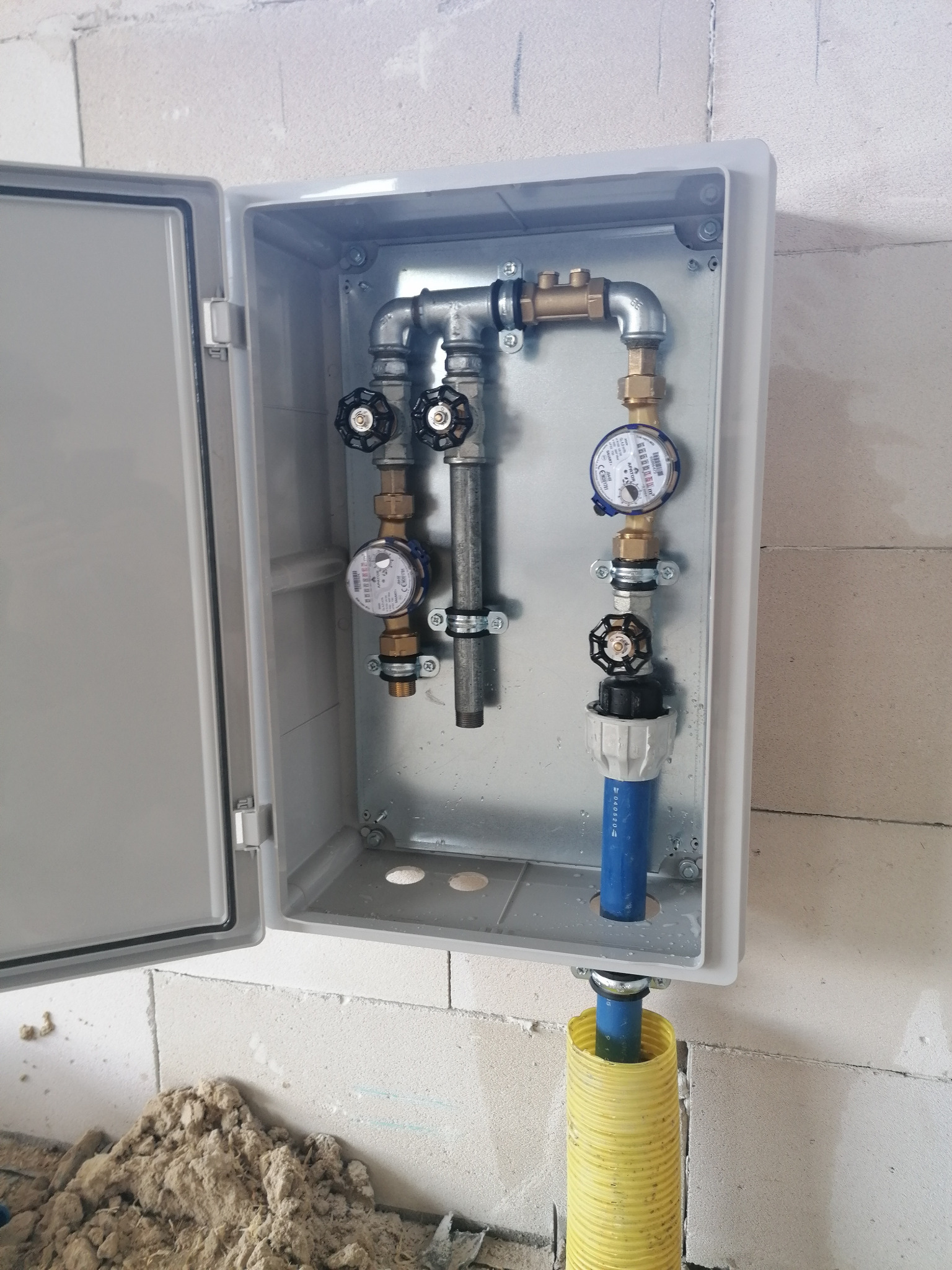 Skrzynka z zaworami i wodomierzami, zamontowana na ścianie z betonu komórkowego, widoczna żółta rura osłonowa.