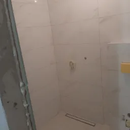 Wykończona kabina prysznicowa z beżowymi płytkami imitującymi marmur, widoczny odpływ liniowy w podłodze i uszczelniona rama kabiny z pianką montażową.