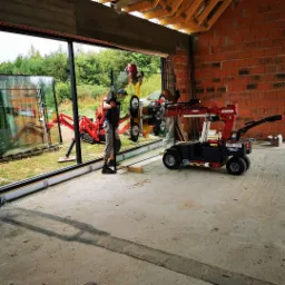 Montaż dużej szyby okiennej za pomocą specjalistycznego podnośnika GW425 na placu budowy, widoczny pracownik w szarych spodniach i czarnej koszulce, w tle surowy mur z cegły i zieleń drzew.