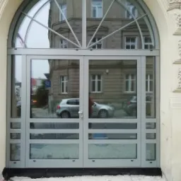 Szklane drzwi zewnętrzne z łukowym nadświetlem, szare ramy, odbicie ulicy i budynków w szybach, widoczne prace wykończeniowe przy wejściu.