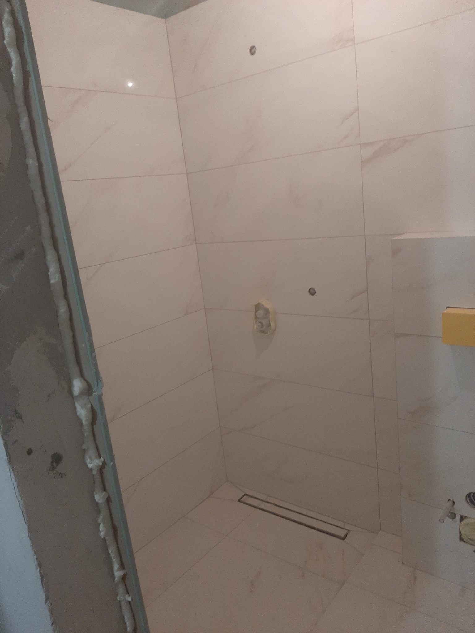 Wykończona kabina prysznicowa z beżowymi płytkami imitującymi marmur, widoczny odpływ liniowy w podłodze i uszczelniona rama kabiny z pianką montażową.