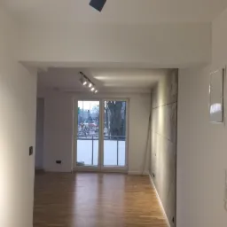 Wnętrze mieszkania po remoncie, z widoczną drewnianą podłogą, ścianą wykończoną panelami imitującymi beton, białymi ścianami i sufitem z oświetleniem punktowym, oraz balkonem z widokiem na zewnątrz.