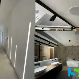 Nowoczesne oświetlenie korytarza z liniowymi lampami LED wpuszczonymi w ścianę, zestawione z oświetleniem sufitowym typu spot w łazience z wanną wolnostojącą i podwójną umywalką.