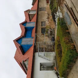 Budynek w trakcie remontu dachu, z częściowo zdjętą starą dachówką i zabezpieczoną folią, widoczne rusztowania i drabina, na ziemi gruz i materiały budowlane.