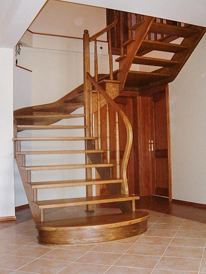 Kręte, drewniane schody wolnostojące z poręczą, prowadzące na wyższą kondygnację, z widocznym fragmentem drzwi i podłogi wyłożonej kafelkami.