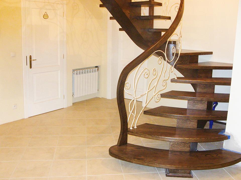 Gięte schody z ciemnego drewna, z ażurową, kremową balustradą, prowadzące na wyższą kondygnację, z widocznymi drzwiami i grzejnikiem w tle.