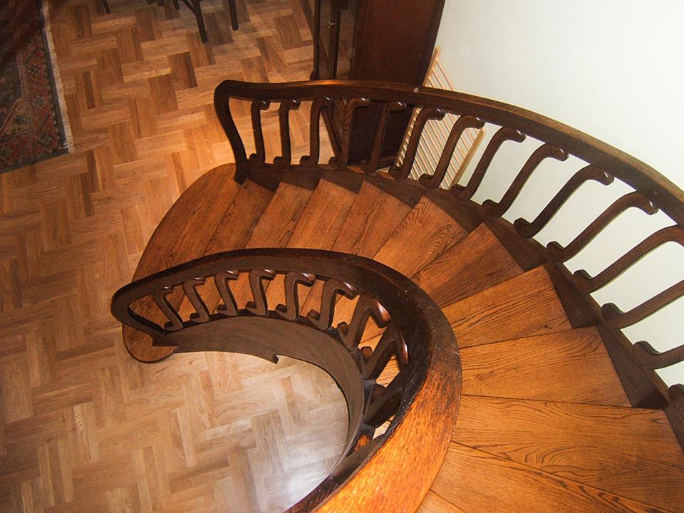 Widok z góry na kręte schody z drewnianymi stopniami i ciemnobrązową, rzeźbioną balustradą, dopasowane do parkietu w jodełkę.