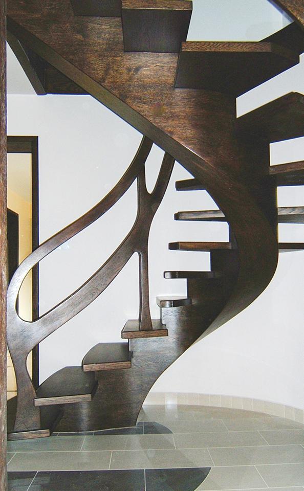 Gięte stopnie z ciemnego drewna, z oryginalną, ażurową balustradą, na tle białej ściany i podłogi z szarych płytek.