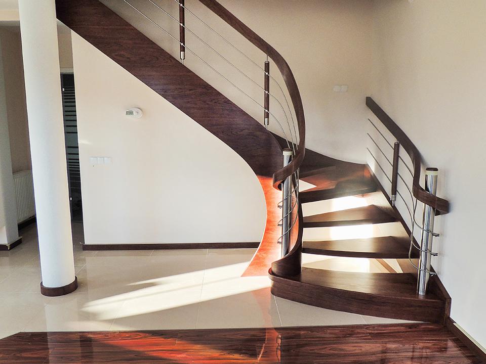 Gięte schody z ciemnego drewna z metalowymi balustradami w nowoczesnym wnętrzu, oświetlone promieniami słońca padającymi na jasną podłogę.