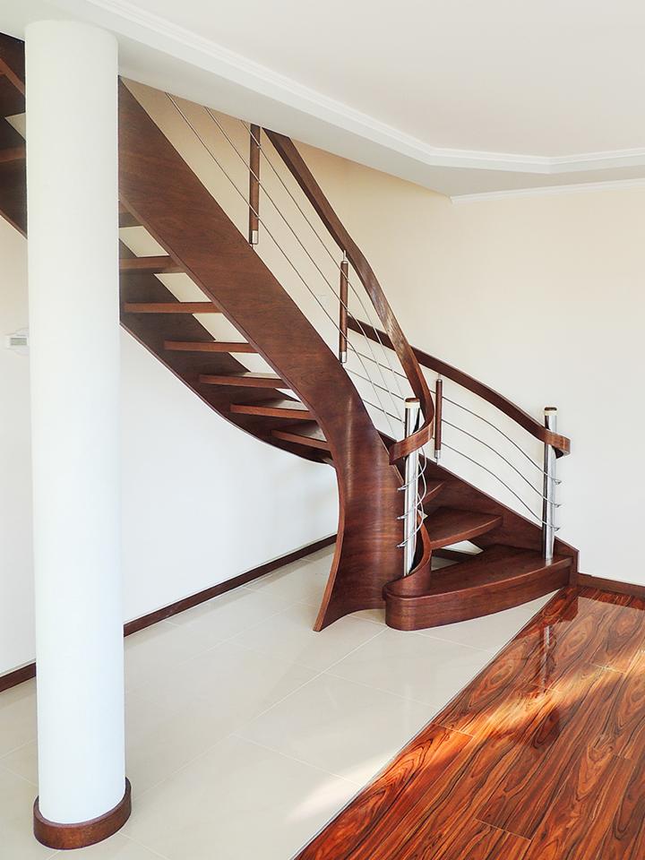 Gięte schody z ciemnego drewna z metalowymi balustradami, widoczne z dołu, z białym filarem po lewej stronie i błyszczącą drewnianą podłogą w tle.