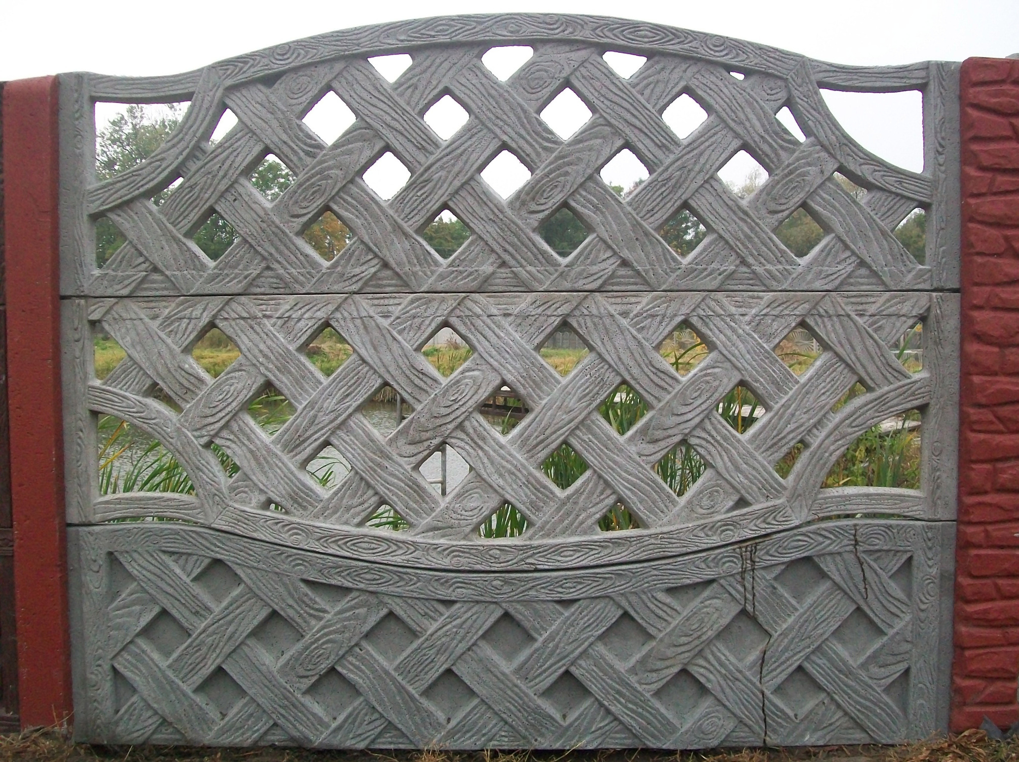 Panel z prefabrykowanego betonu imitującego drewno, tworzący ażurowe ogrodzenie z czerwonymi słupkami, widok na zewnątrz przez romboidalne prześwity.