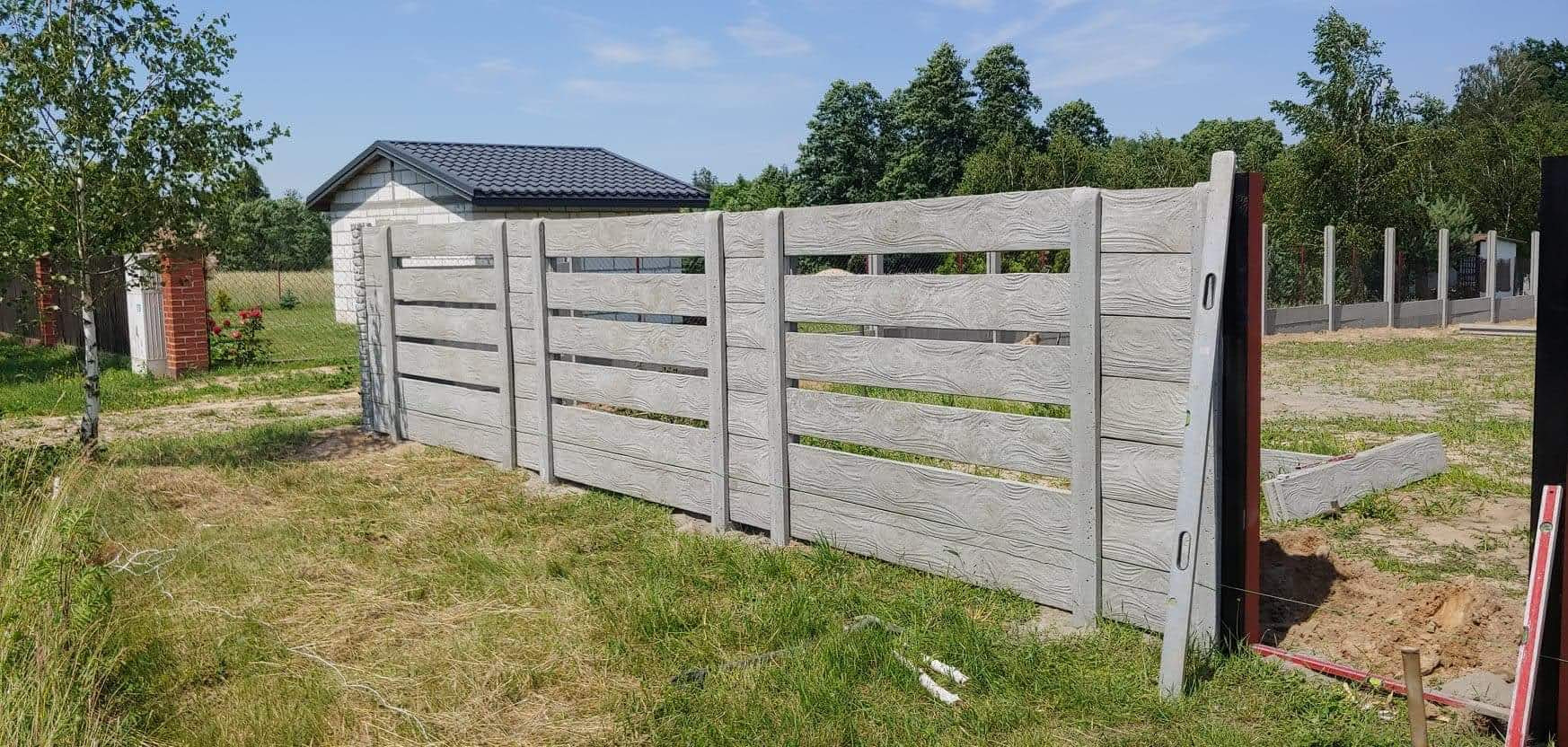 Fragment betonowego ogrodzenia z poziomymi panelami imitującymi drewno, widoczne poziomice i niedokończone elementy w trawie, w tle dom z szarą dachówką i drzewa.