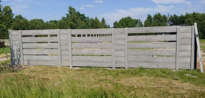 Fragment nowego, szarego ogrodzenia betonowego z poziomymi szczelinami, imitującego drewno, częściowo opartego o ziemię, z poziomicą opartą o jeden z paneli.