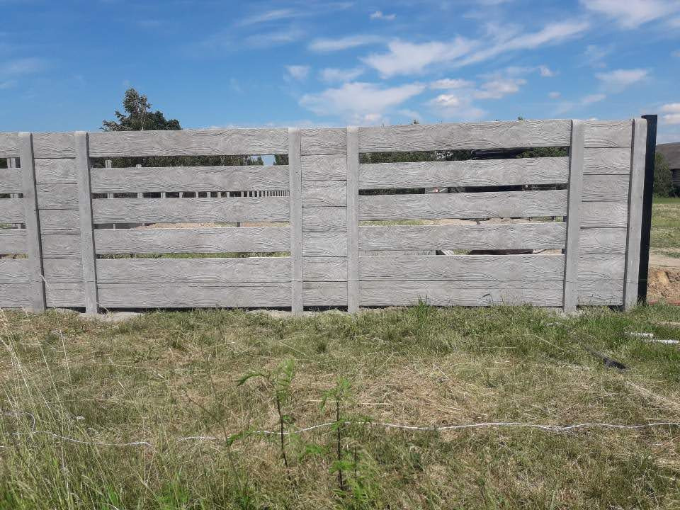 Nowo postawione, prefabrykowane, betonowe przęsła ogrodzeniowe imitujące drewno, z widocznymi szczelinami między poziomymi elementami, na tle trawy i błękitnego nieba.