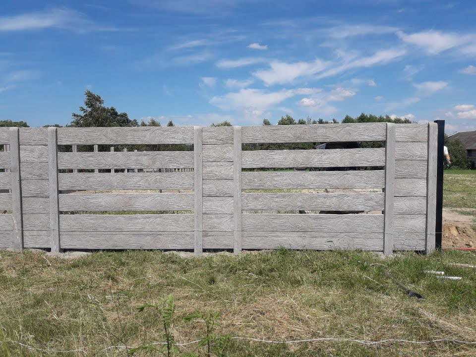 Szare, betonowe przęsła ogrodzeniowe z fakturą drewna, widoczne na tle błękitnego nieba i trawy, z czarnym słupkiem na skraju.