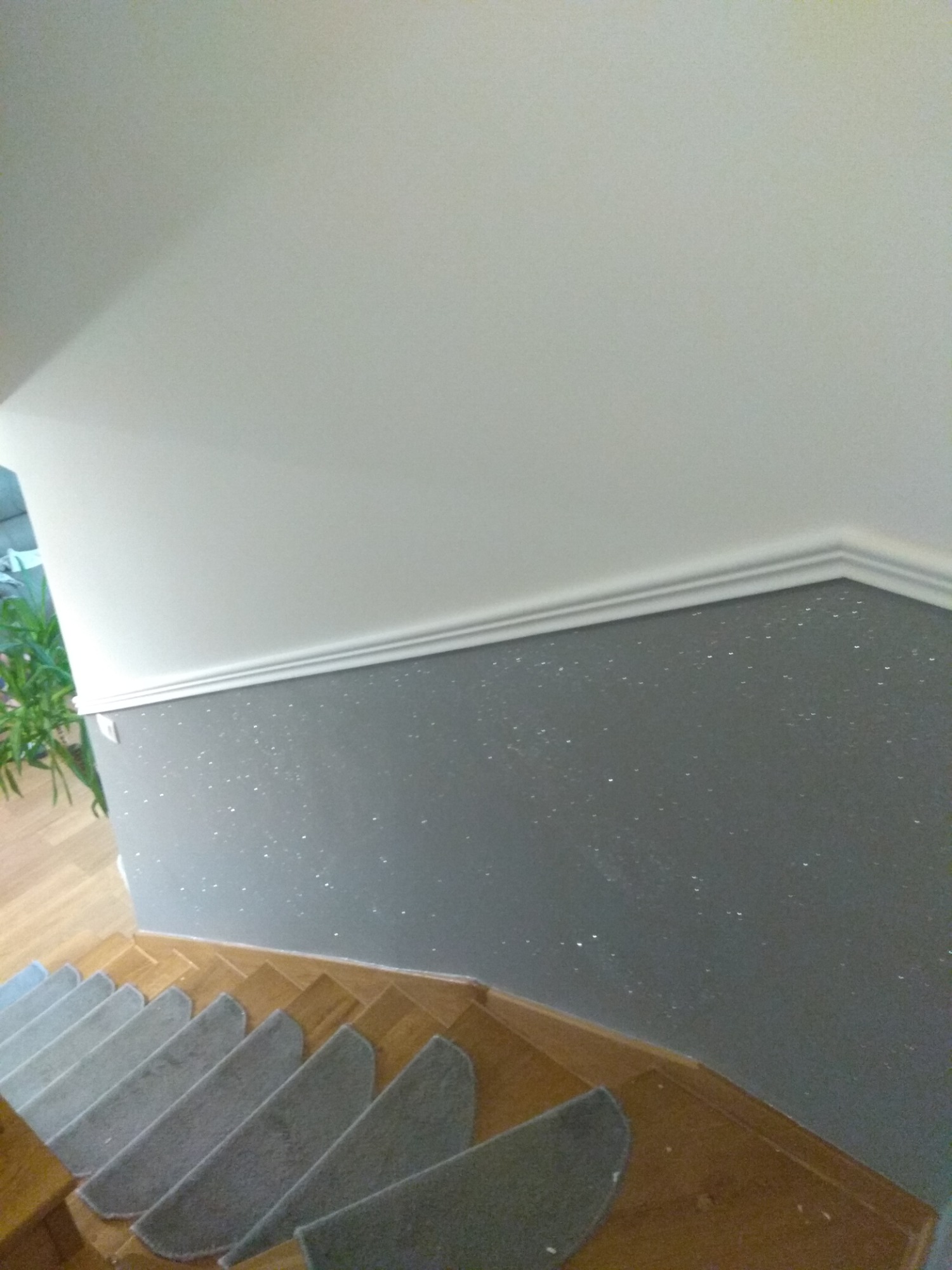 Szara ściana ze srebrnymi drobinkami na klatce schodowej, biały sufit z ozdobnym gzymsem, drewniane schody z szarymi nakładkami antypoślizgowymi.