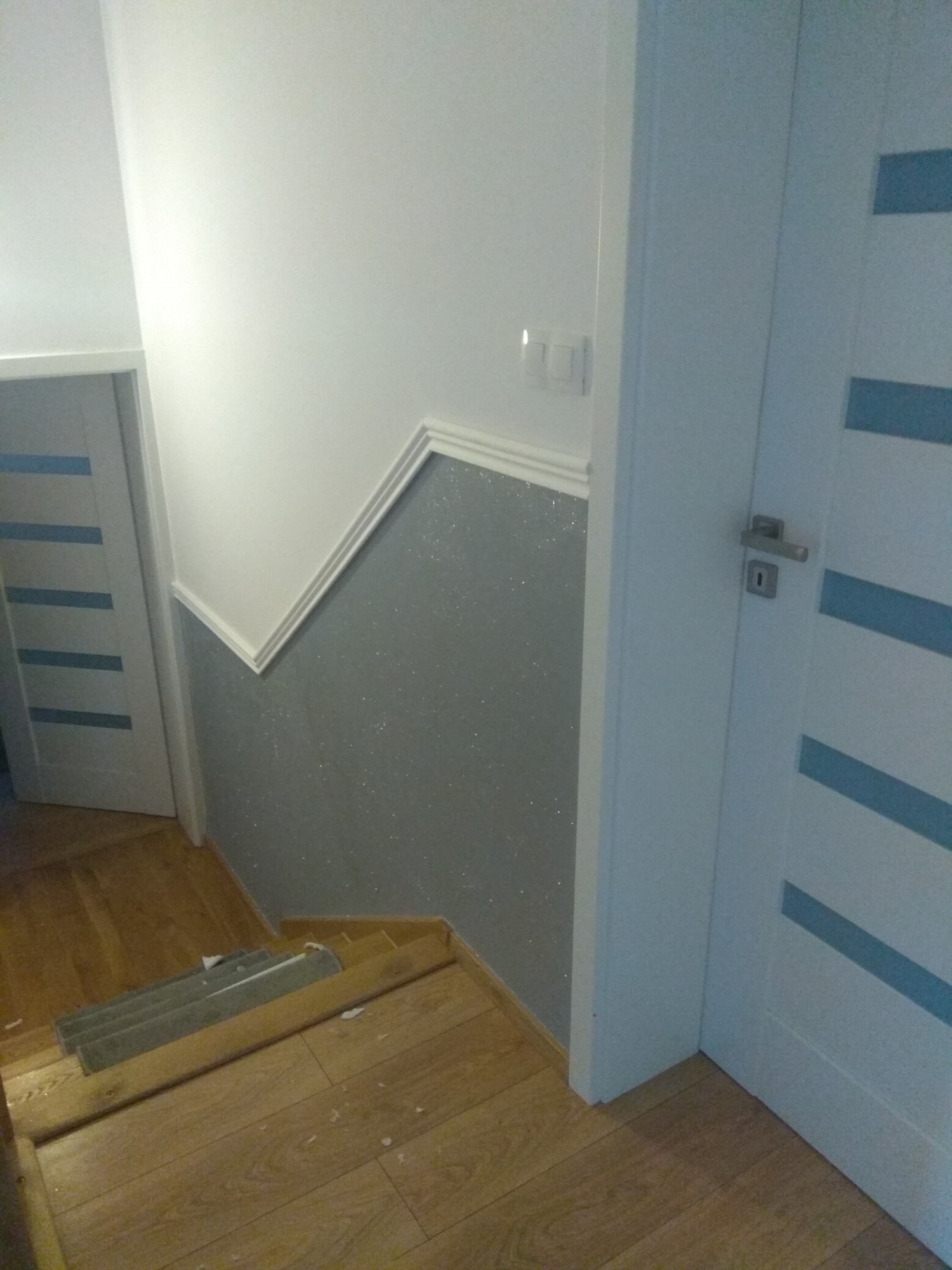 Klatka schodowa z pomalowaną na szaro dolną częścią ściany z efektem brokatu, białą górną częścią oddzieloną listwą dekoracyjną, drewnianymi schodami i białymi drzwiami z poziomymi, niebieskimi...