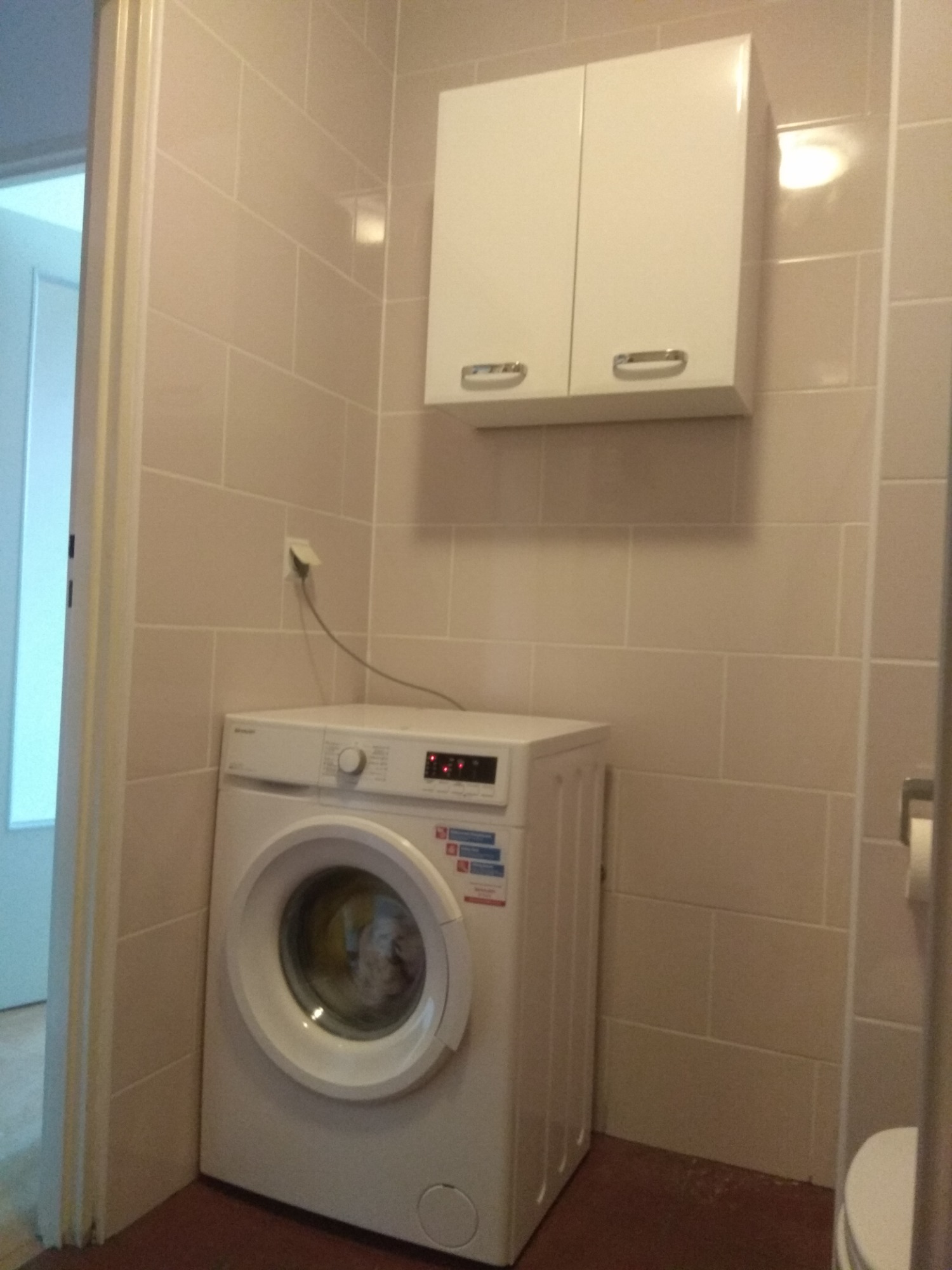 Mała łazienka z beżowymi płytkami, białą pralką i szafką wiszącą. Widoczny fragment otwartych drzwi do innego pomieszczenia.