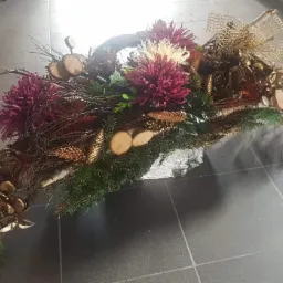 Kompozycja kwiatowa z bordowymi daliami, złotymi szyszkami, drewnianymi krążkami i zielonymi gałązkami na korze brzozy, ozdobiona złotą siatką.