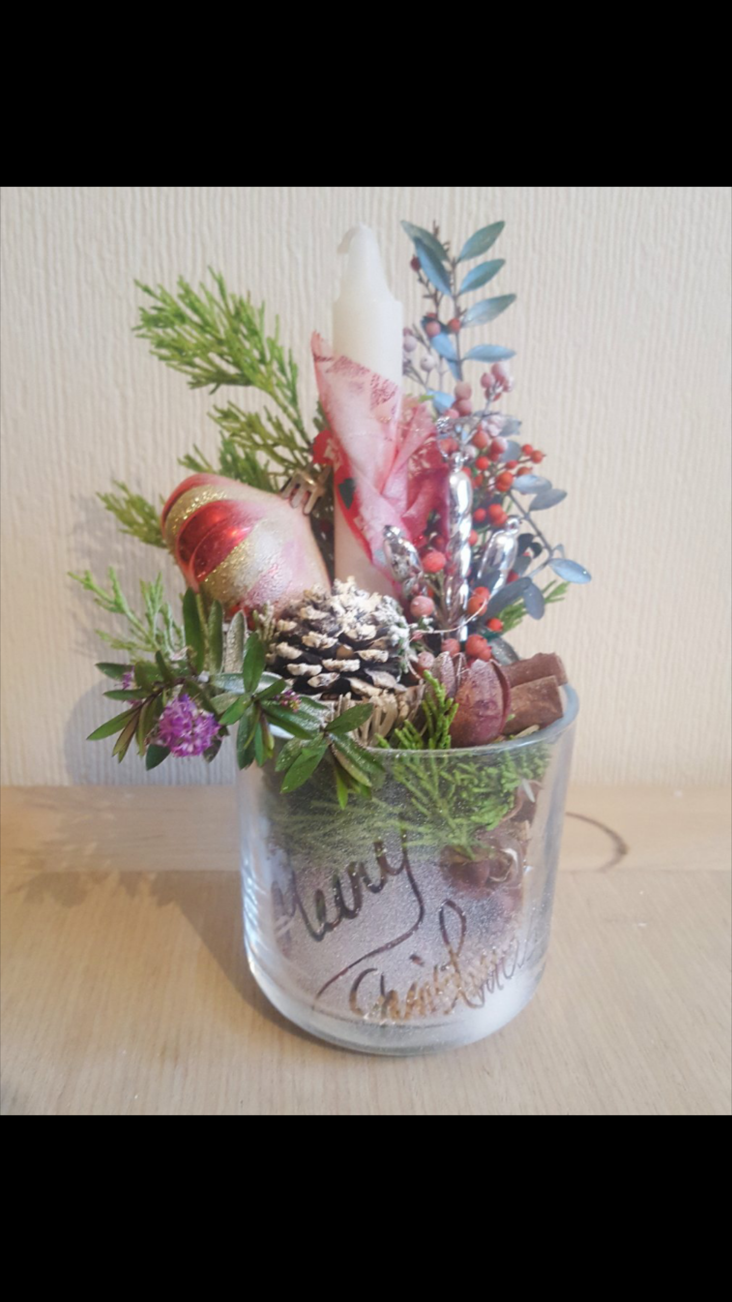 Świąteczna kompozycja w szklanym naczyniu z napisem 'Merry Christmas', zawierająca świecę, bombkę, szyszki, gałązki świerku i inne dekoracje.