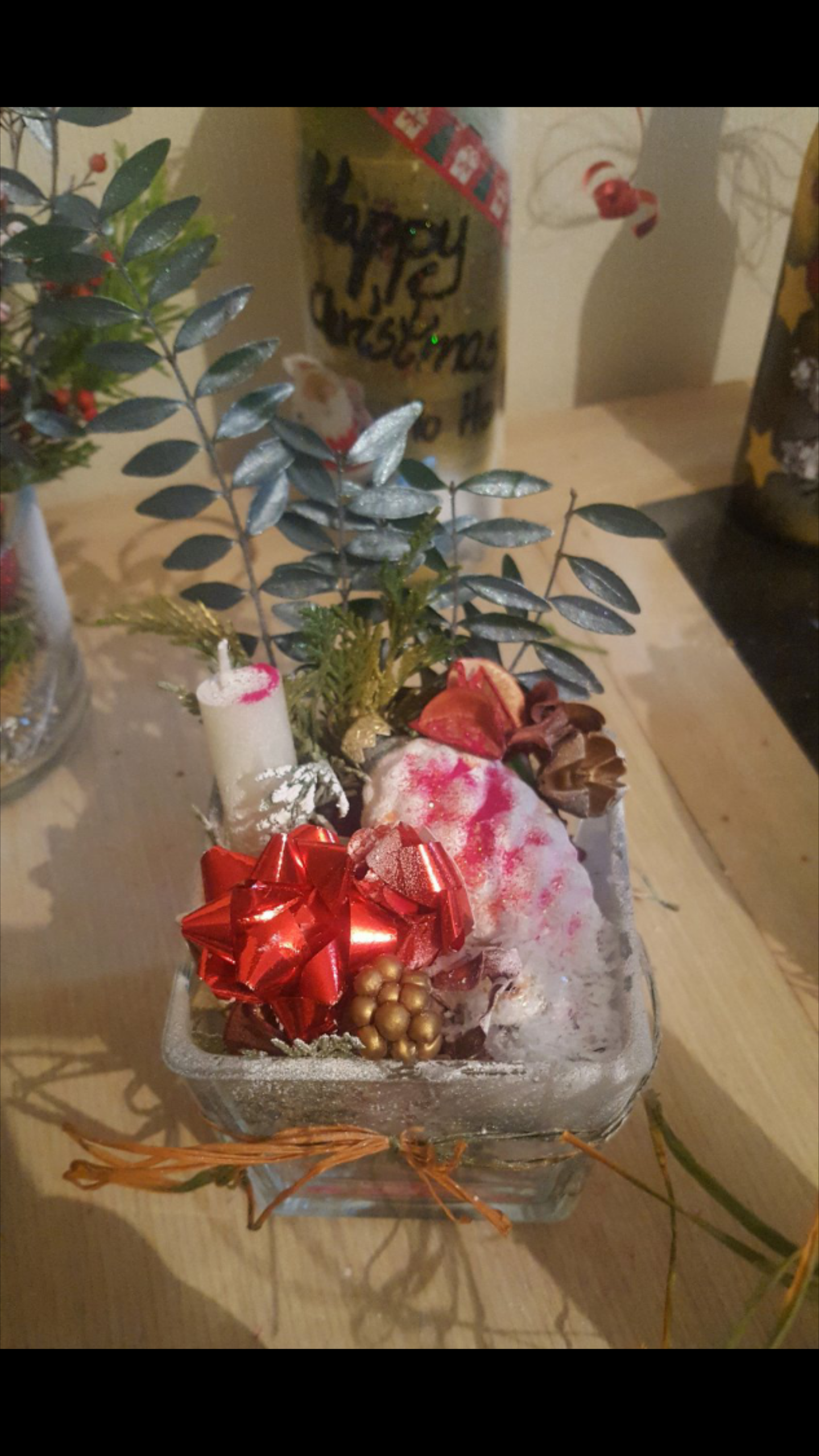 Dekoracja świąteczna w szklanym naczyniu z czerwoną kokardą, złotymi kulkami, sztucznym śniegiem i świeczką, na drewnianym blacie, z elementami dekoracyjnymi w tle.